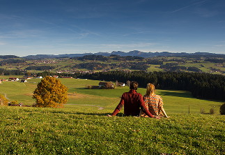Paar sitzt auf einer Wiese und blickt auf eine malerische Landschaft mit Bergen und Wäldern im Hintergrund, ideal für romantische Ausflüge in der Natur.