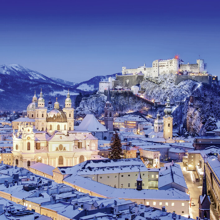Salzburg bei Nacht mit verschneiten Dächern, der Festung Hohensalzburg im Hintergrund und einer malerischen Winterlandschaft.