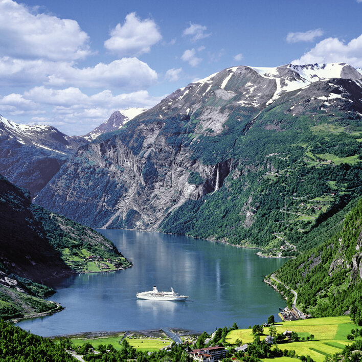 Blick auf den Geirangerfjord mit einem Kreuzfahrtschiff, umgeben von majestätischen Bergen und grünen Wiesen.