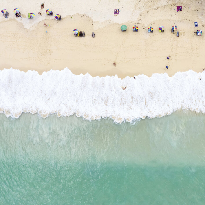 Strandansicht mit Sonnenschirmen, Liegen und Wellen im türkisfarbenen Wasser.