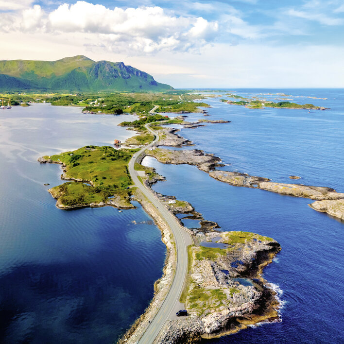 Luftaufnahme einer malerischen Küstenstraße, die durch grüne Landschaften und über felsige Ufer in Norwegen führt.