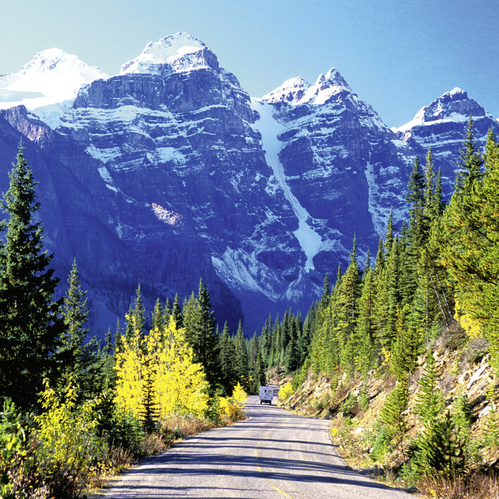 Berglandschaft mit schneebedeckten Gipfeln und einer kurvenreichen Straße, gesäumt von grünen und gelben Bäumen.